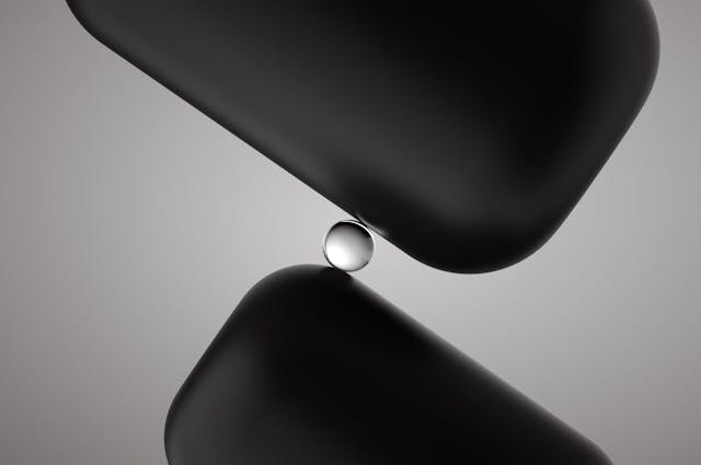 Image of a small glass sphere balancing between two big smooth cubes on grey background