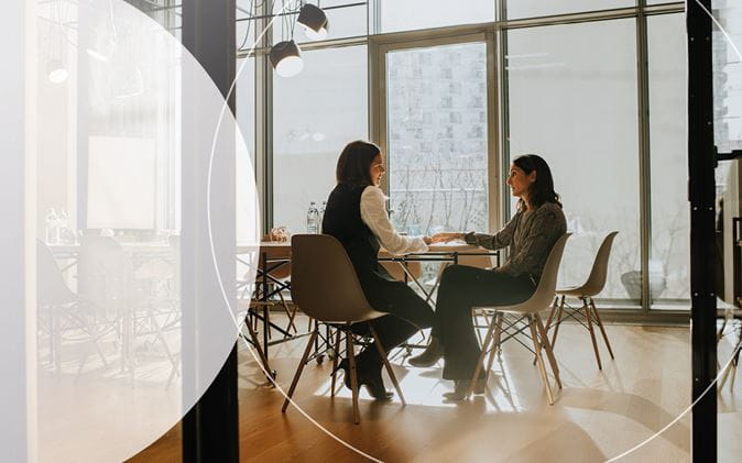 Two women sitting at a boardroom table, floor to table windows and sunlight coming in