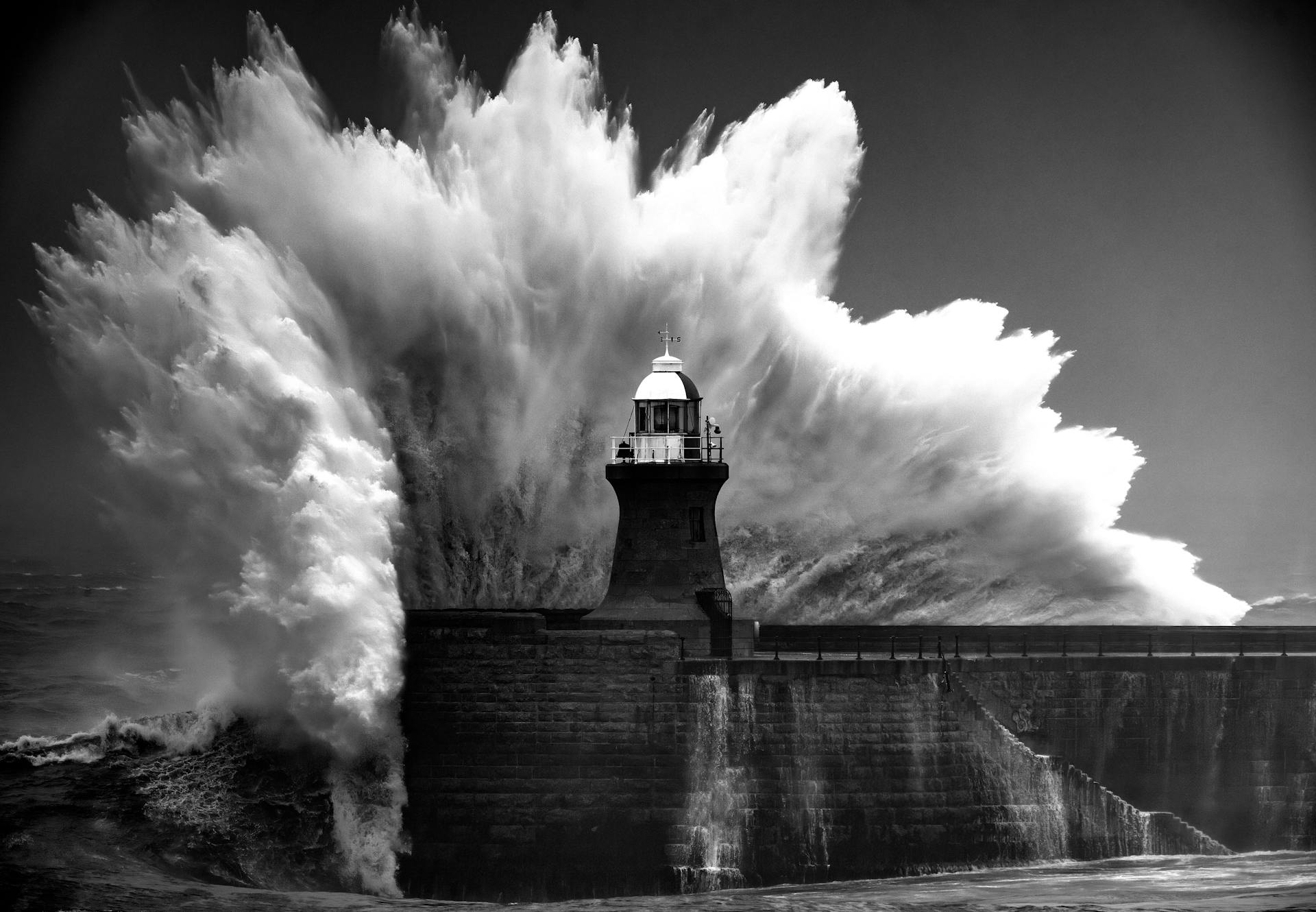 A huge wave rises behind a pier and a lighthouse in a black and white image