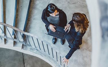 Two colleagues standing on a stair case talking, one holding laptop.