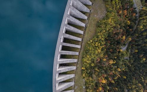 Ariel shot of a large water dam on left, with green trees beneath on the right