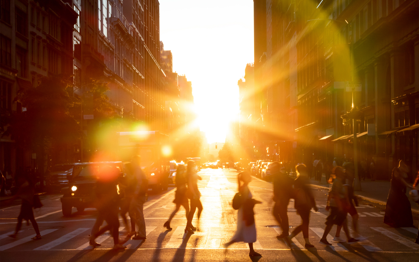 The sun reflecting against buildings as people walk across a busy street