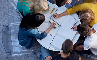Group of people working at a circular desk together, writing on paper documents. 