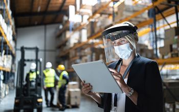 Manager with protective shield using tablet indoors in warehouse, coronavirus concept
