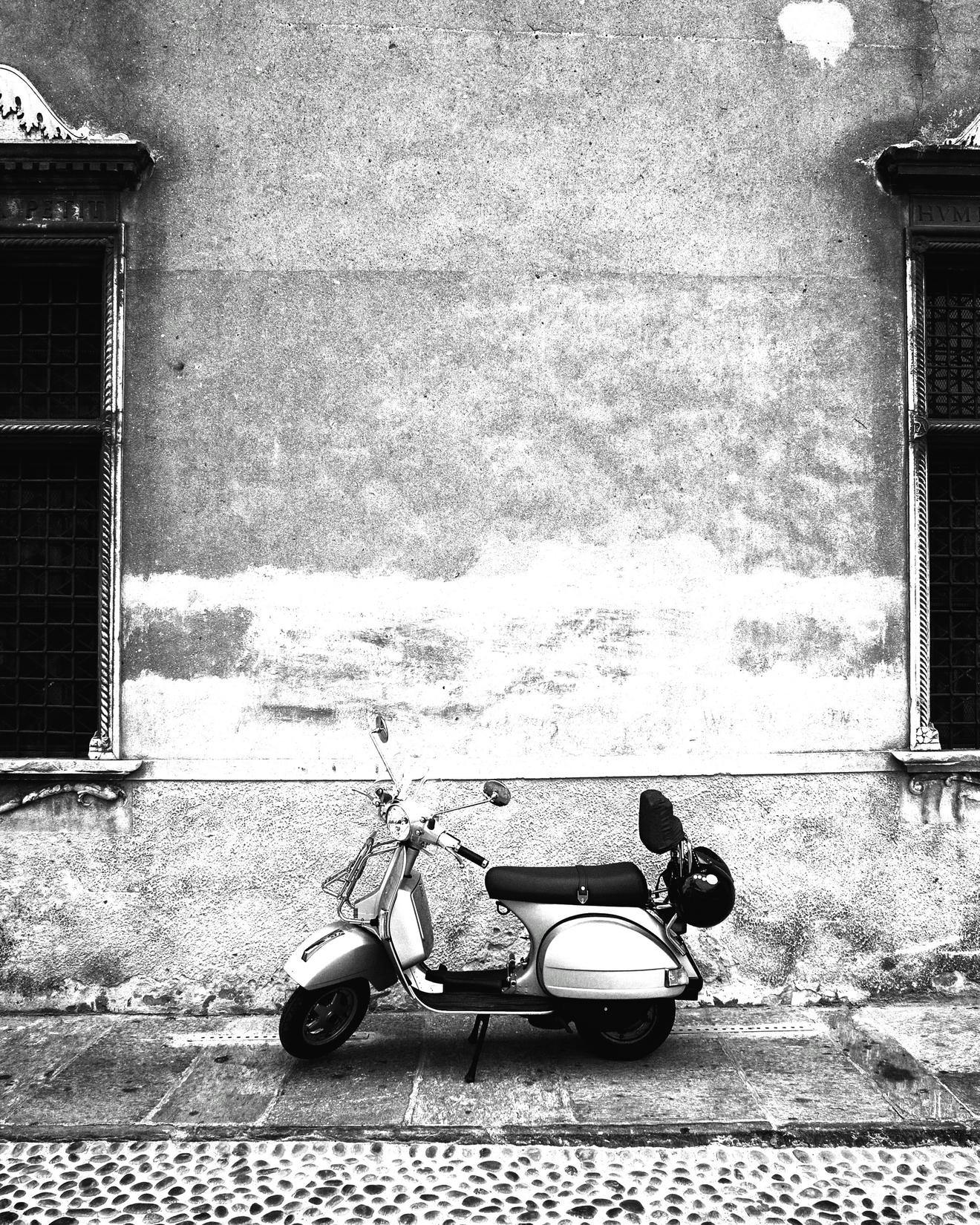 Black and white image of a Vespa scooter against a wall with ornate windows in Italy